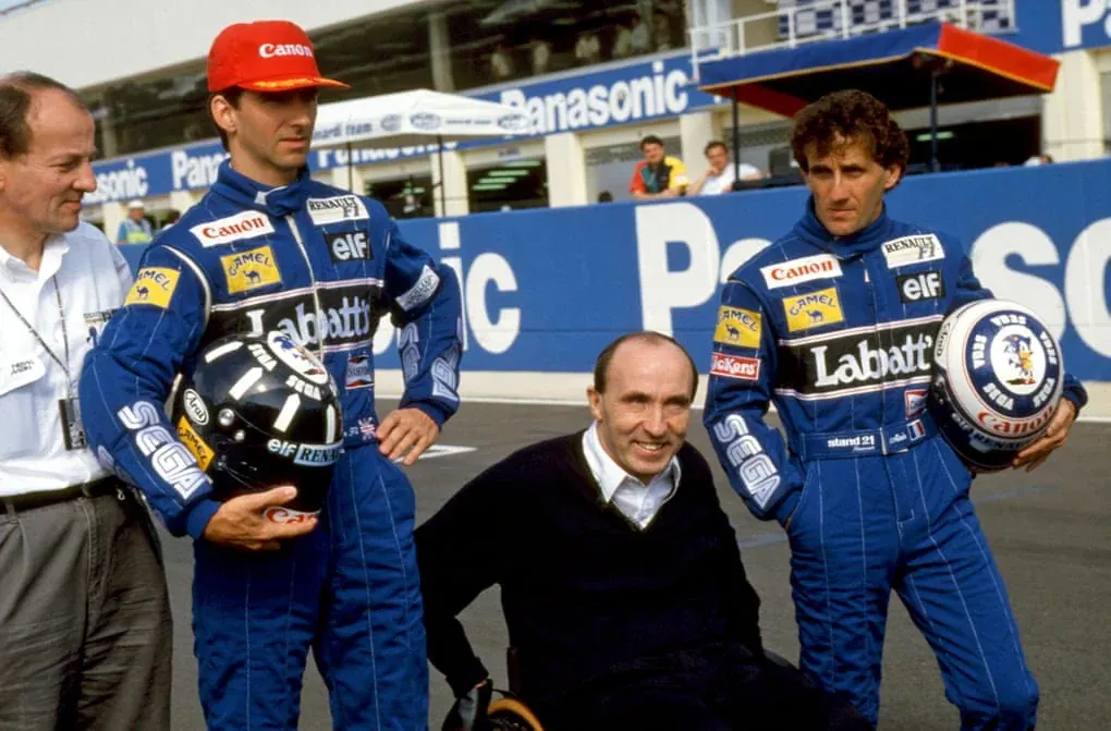 Frank Williams smiling between Alain Prost and Damon Hill, both holding helmets, in 1993.