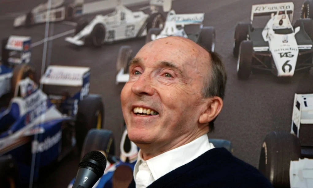 Frank Williams laughing, with photographs of Williams racing cars behind him in 2013.