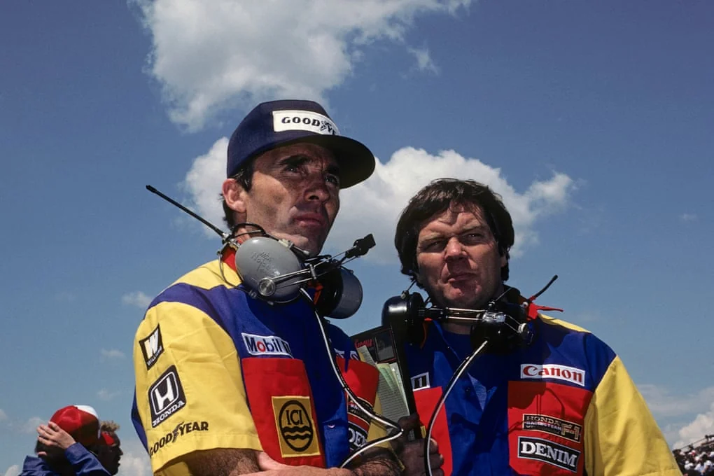 Frank Williams and Patrick Head stand together wearing team jackets and headsets in 1985.