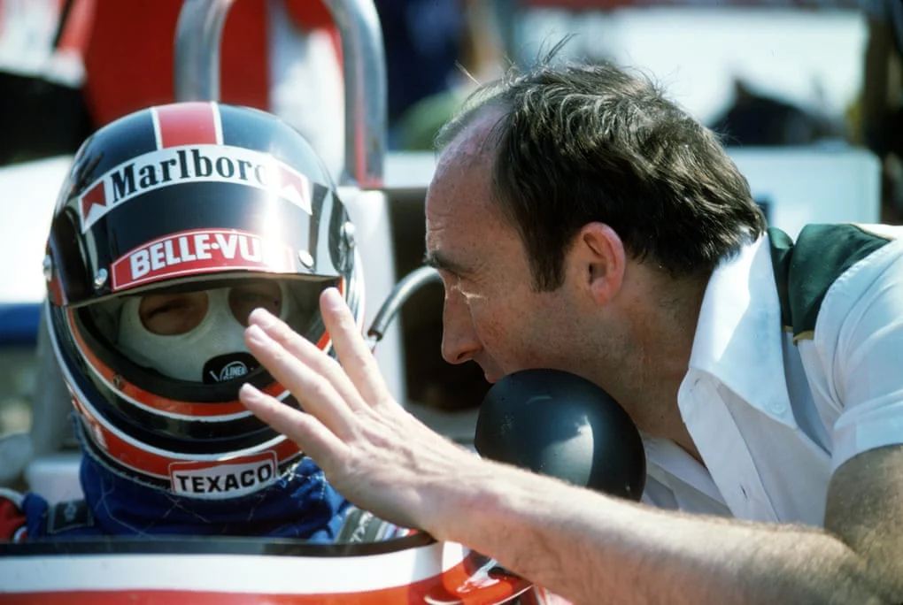 Frank Williams leans toward a helmeted driver in the cockpit at Monza in 1977.