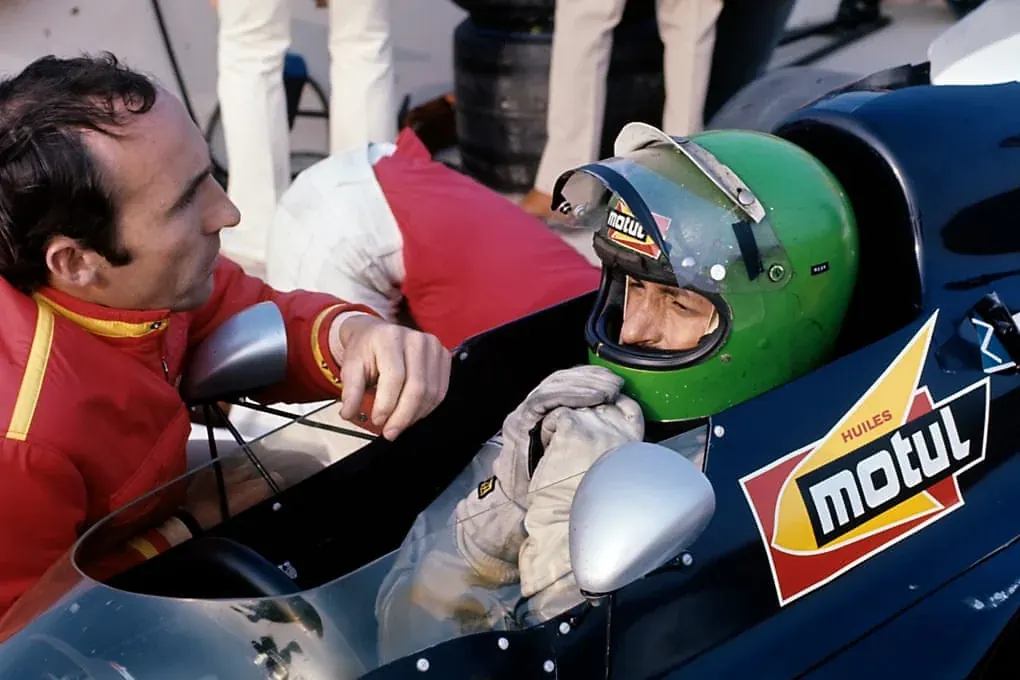 Frank Williams talks closely with Henri Pescarolo seated in a blue racing car in 1971.