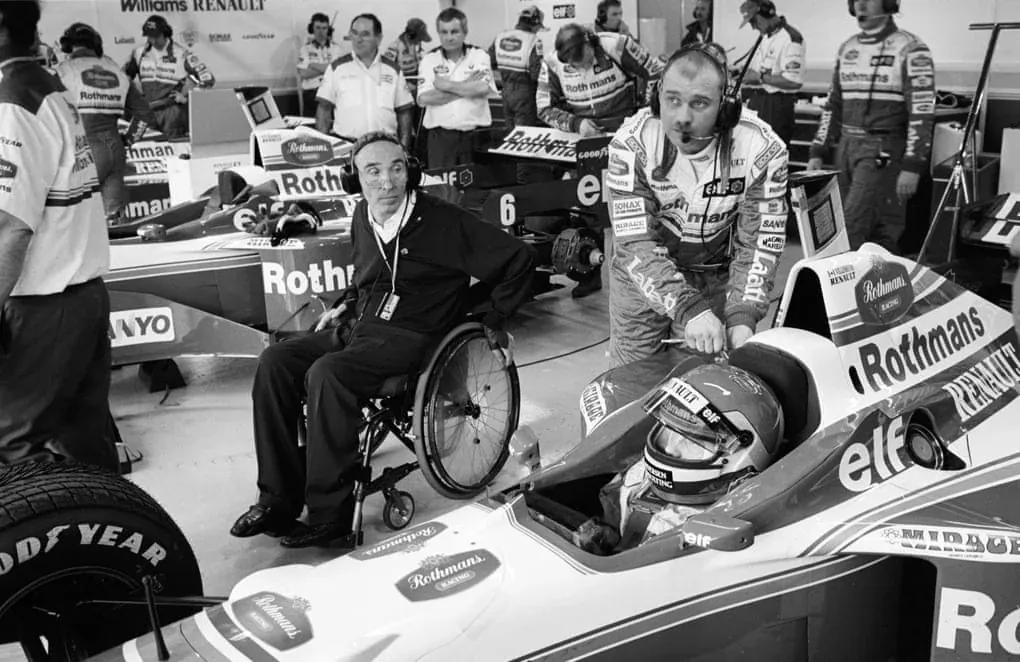 Frank Williams beside Jacques Villeneuve and a Williams car, surrounded by the team in 1996.